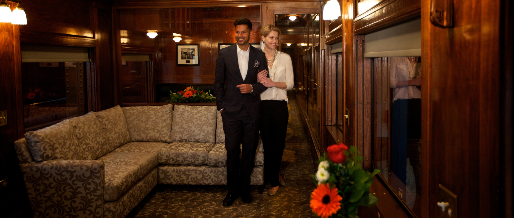 A man and a woman stand in a lounging area inside of a rail car.