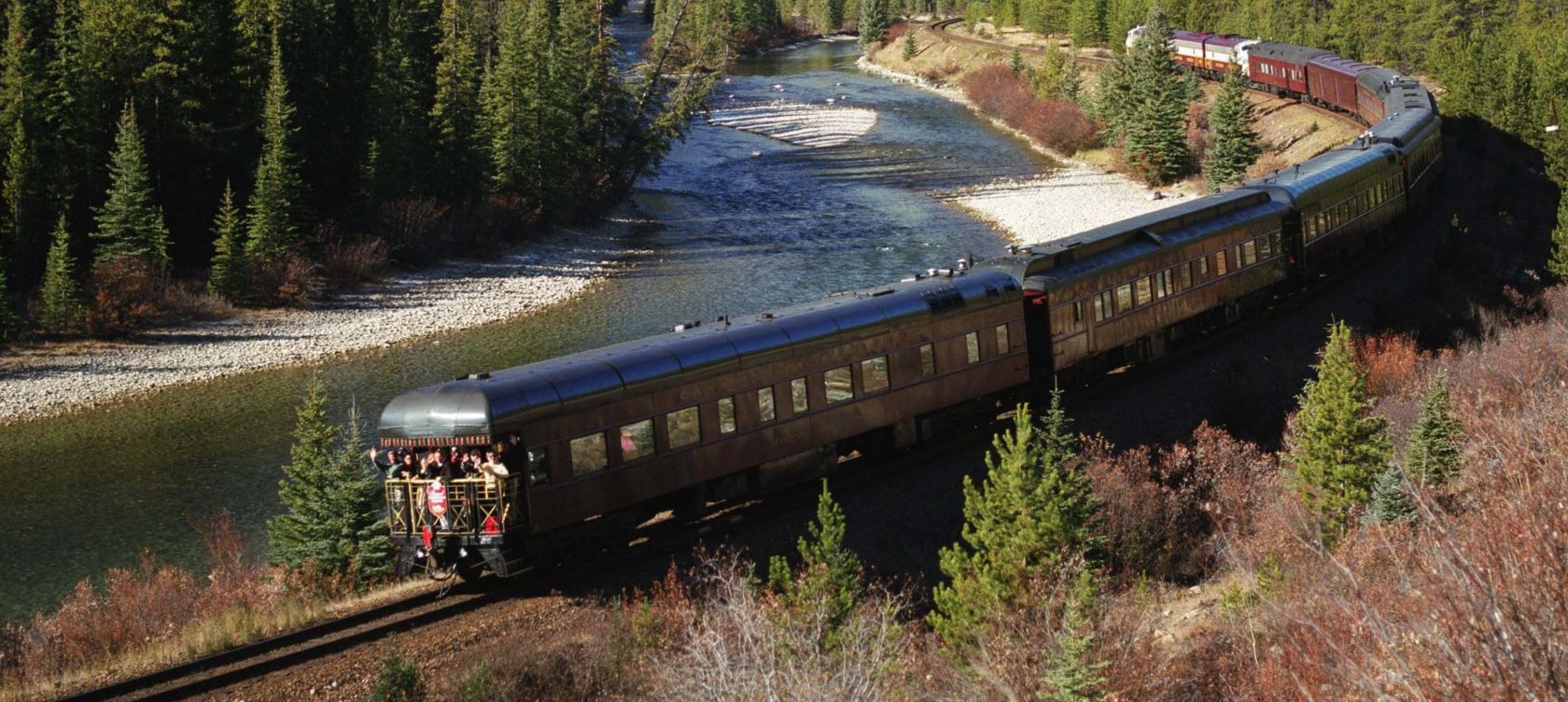 A train moving alongside a river and a forest.